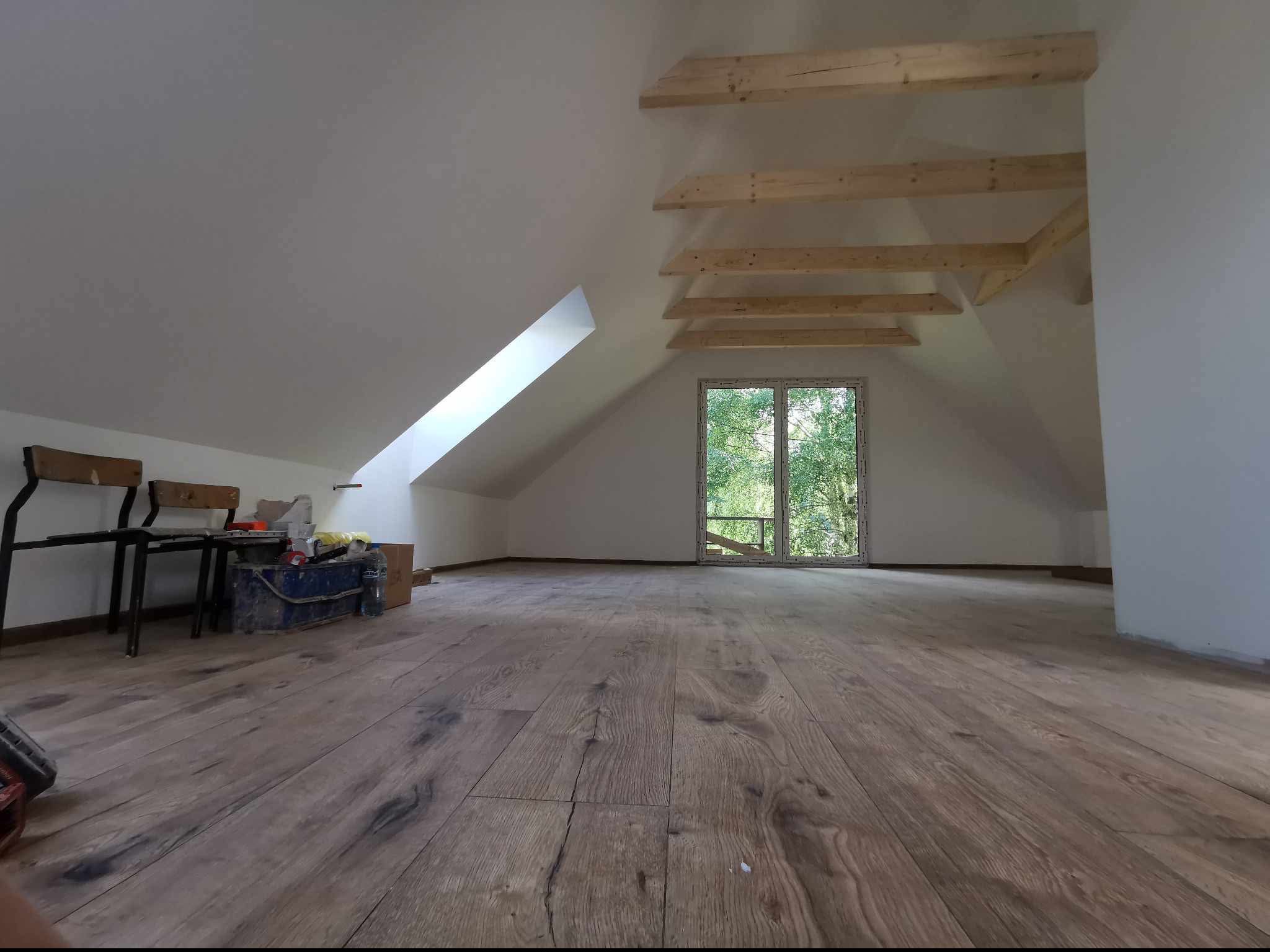 Poddasze po remoncie z drewnianą podłogą, widoczne narzędzia i krzesła w tle, okno dachowe z widokiem na drzewa.