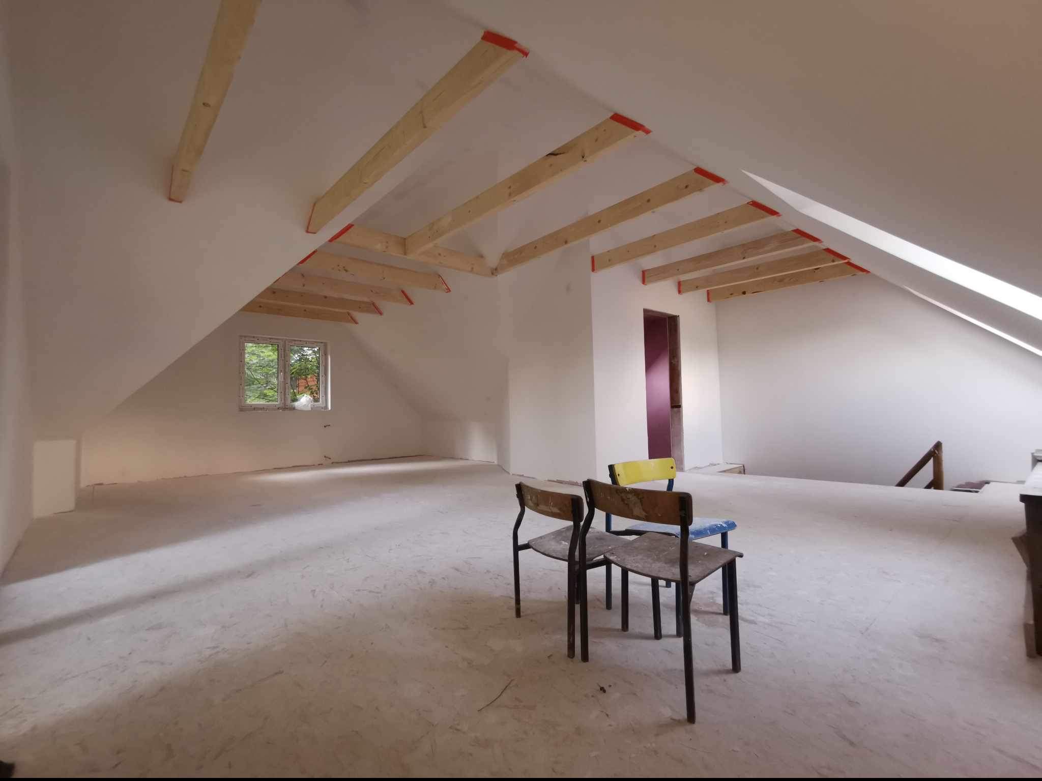Poddasze w trakcie adaptacji, widoczne drewniane belki stropowe z pomarańczową taśmą zabezpieczającą, białe ściany i podłoga, okno z widokiem na zieleń oraz trzy krzesła w centrum pomieszczenia.