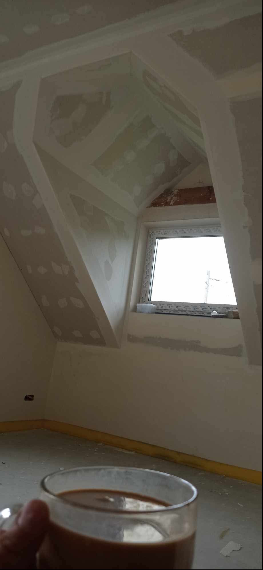 Poddasze w trakcie wykańczania: płyty karton-gips na skosach, szpachlowane łączenia, okno dachowe, kubek z kawą w dolnej części kadru.