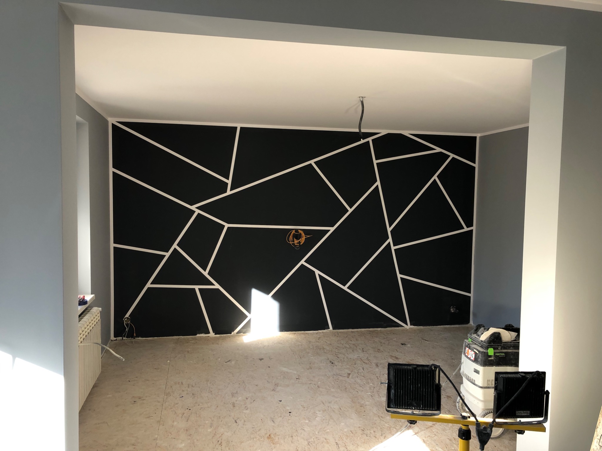 Pokój w trakcie remontu z pomalowaną na czarno ścianą, na której znajdują się białe, geometryczne wzory. Widoczne narzędzia i lampy budowlane, surowe podłoże i grzejnik.