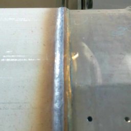 SPAWAMY NA JAKOŚĆ Marcin Dębiński - Spoina na metalowej rurze, widoczne przebarwienia termiczne i otwory montażowe, w tle elementy konstrukcyjne hali.