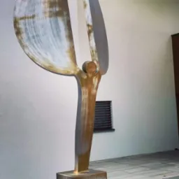 Abstrakcyjna rzeźba metalowa przedstawiająca stylizowaną postać z rozpostartymi ramionami i dużymi, półkolistymi elementami przypominającymi skrzydła, umieszczona na cokole przed białą ścianą z oknem.