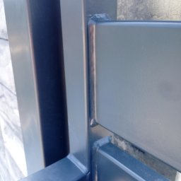 SPAWAMY NA JAKOŚĆ Marcin Dębiński - Fragment konstrukcji metalowej, widoczne spawy łączące elementy, szlifowane i pomalowane na ciemny kolor, z lekkim połyskiem, na tle betonowej ściany.