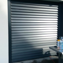 SPAWAMY NA JAKOŚĆ Marcin Dębiński - Szare, poziome żaluzje zewnętrzne zamontowane na ścianie budynku, widoczne z bliska, z elementami wyposażenia na stole z boku.