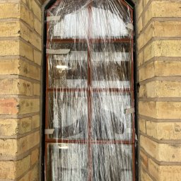 SPAWAMY NA JAKOŚĆ Marcin Dębiński - Metalowa konstrukcja okienna z łukowym nadprożem, zabezpieczona folią i taśmą, w surowym murze z cegły klinkierowej w odcieniach beżu i czerwieni.