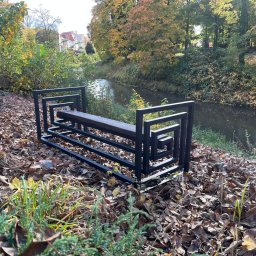 SPAWAMY NA JAKOŚĆ Marcin Dębiński - Nowoczesna, metalowa ławka parkowa z drewnianym siedziskiem, o geometrycznym designie, ustawiona na tle jesiennego krajobrazu z rzeką i drzewami.