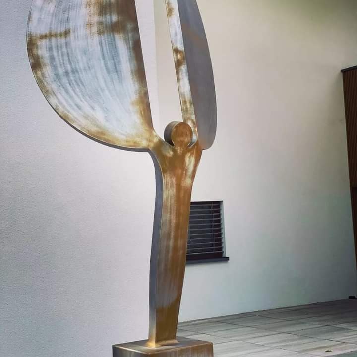 Abstrakcyjna rzeźba metalowa przedstawiająca stylizowaną postać z rozpostartymi ramionami i dużymi, półkolistymi elementami przypominającymi skrzydła, umieszczona na cokole przed białą ścianą z oknem.