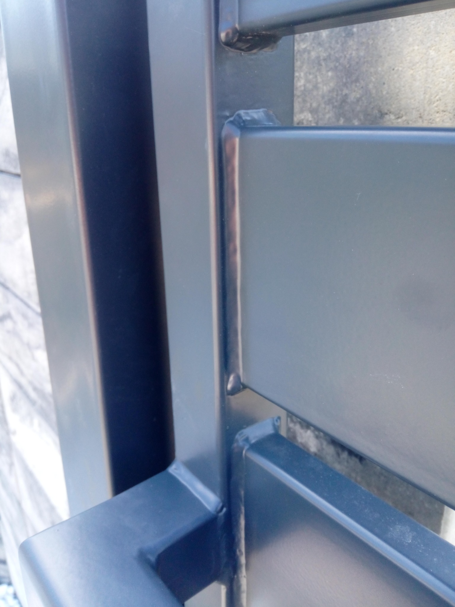 Fragment konstrukcji metalowej, widoczne spawy łączące elementy, szlifowane i pomalowane na ciemny kolor, z lekkim połyskiem, na tle betonowej ściany.