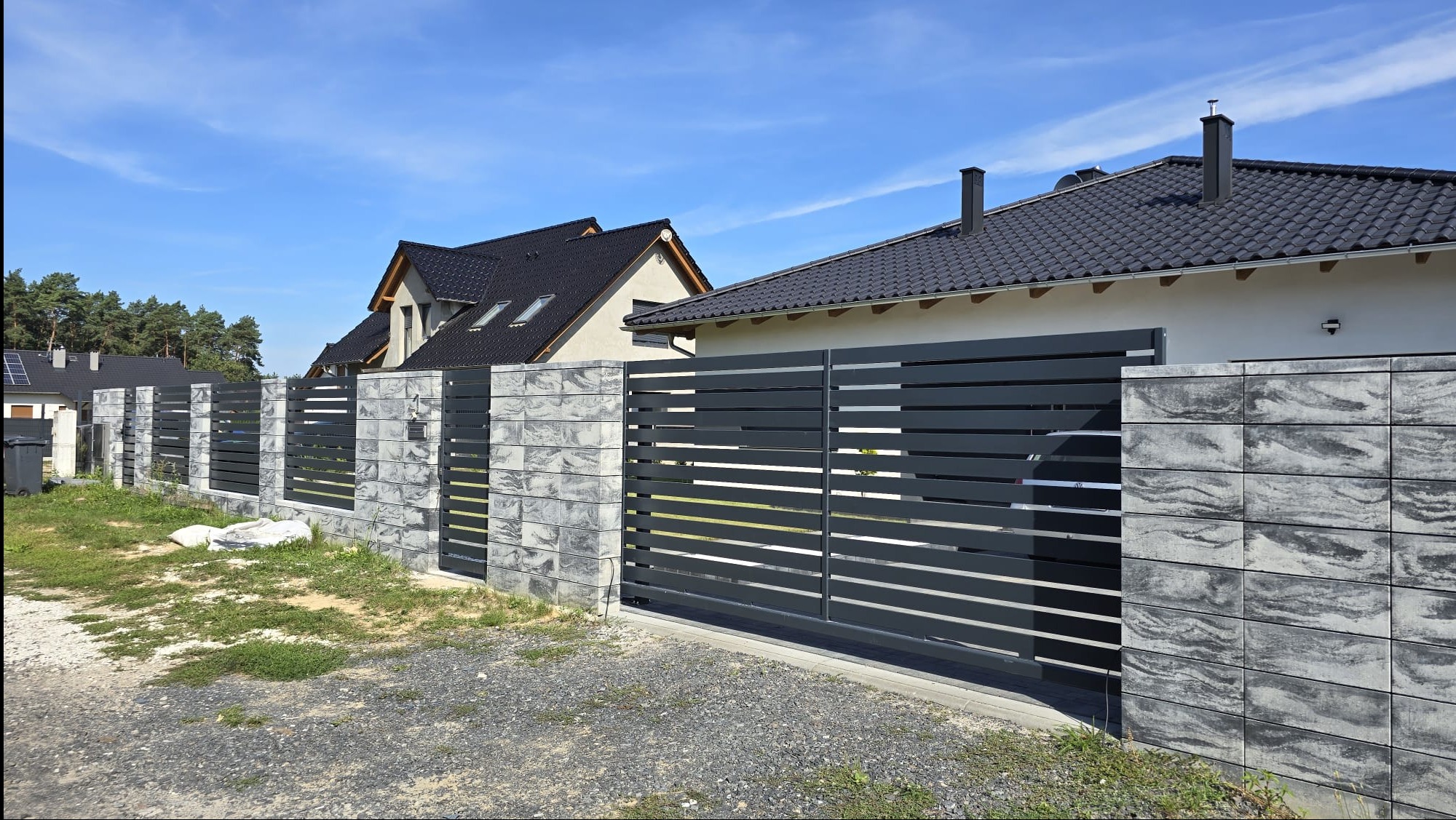 Nowoczesna brama przesuwna w kolorze grafitowym z poziomymi szczeblami, osadzona w murze z szarej, wzorzystej kostki, na tle domów jednorodzinnych i błękitnego nieba.