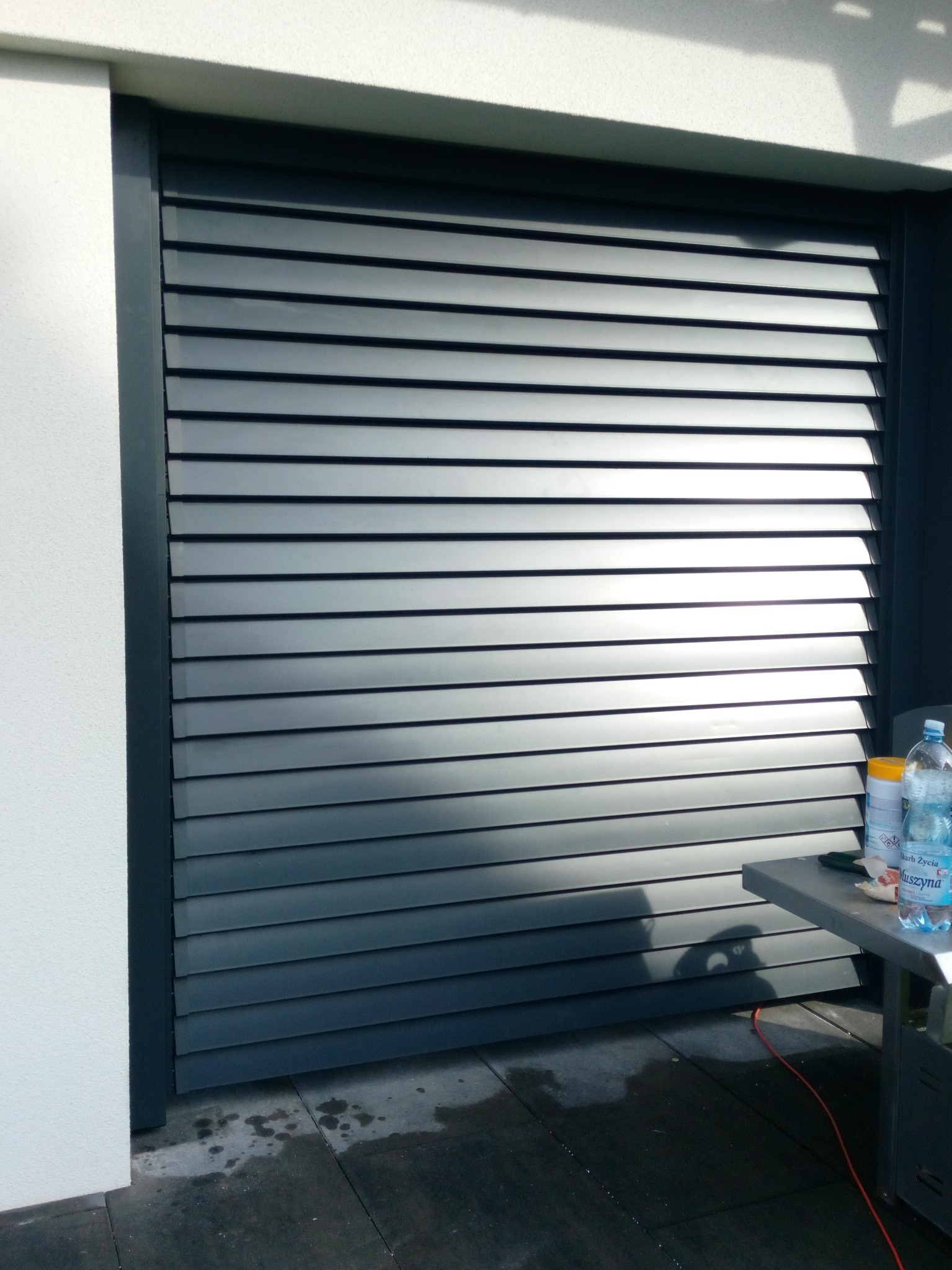 Szare, poziome żaluzje zewnętrzne zamontowane na ścianie budynku, widoczne z bliska, z elementami wyposażenia na stole z boku.