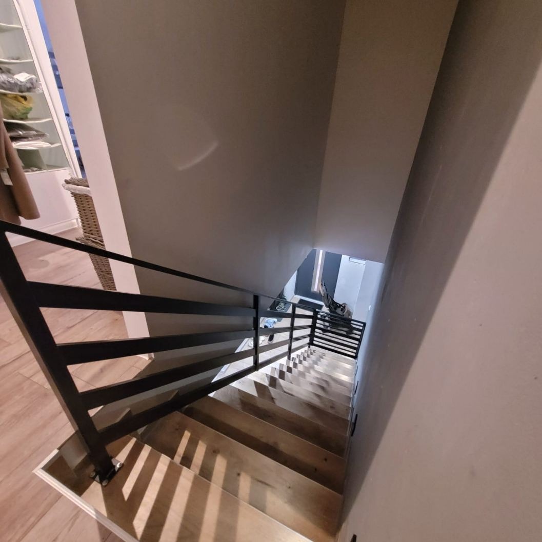 Widok z góry na drewniane schody z czarną, minimalistyczną balustradą, rzucającą cienie na stopnie, z widocznym fragmentem pomieszczenia na górze i dole schodów.