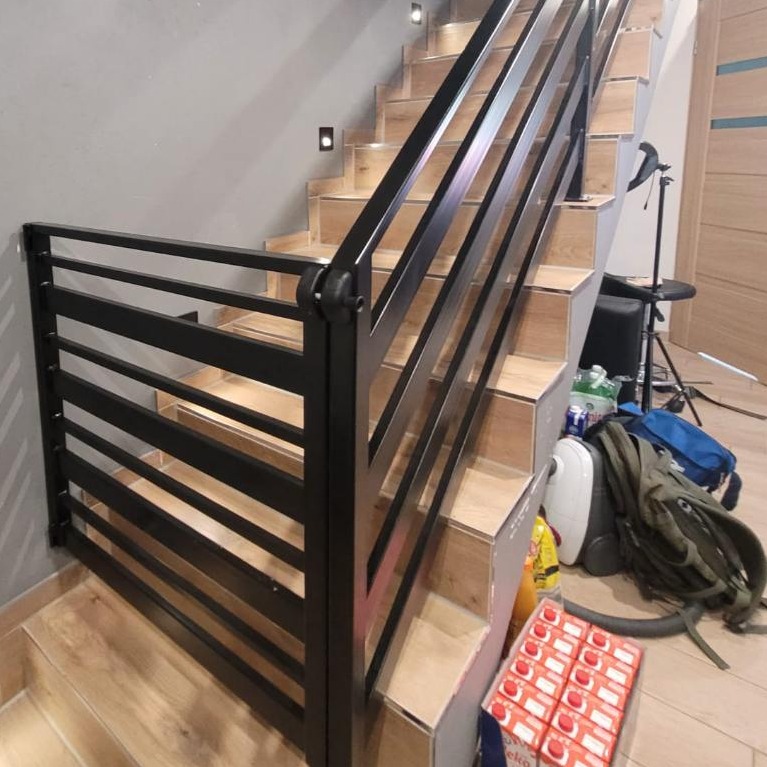 Czarna, pozioma balustrada schodowa z metalowych profili na drewnopodobnych schodach, widok z dołu na bieg schodów, z widocznymi elementami wyposażenia i materiałów budowlanych na schodach.