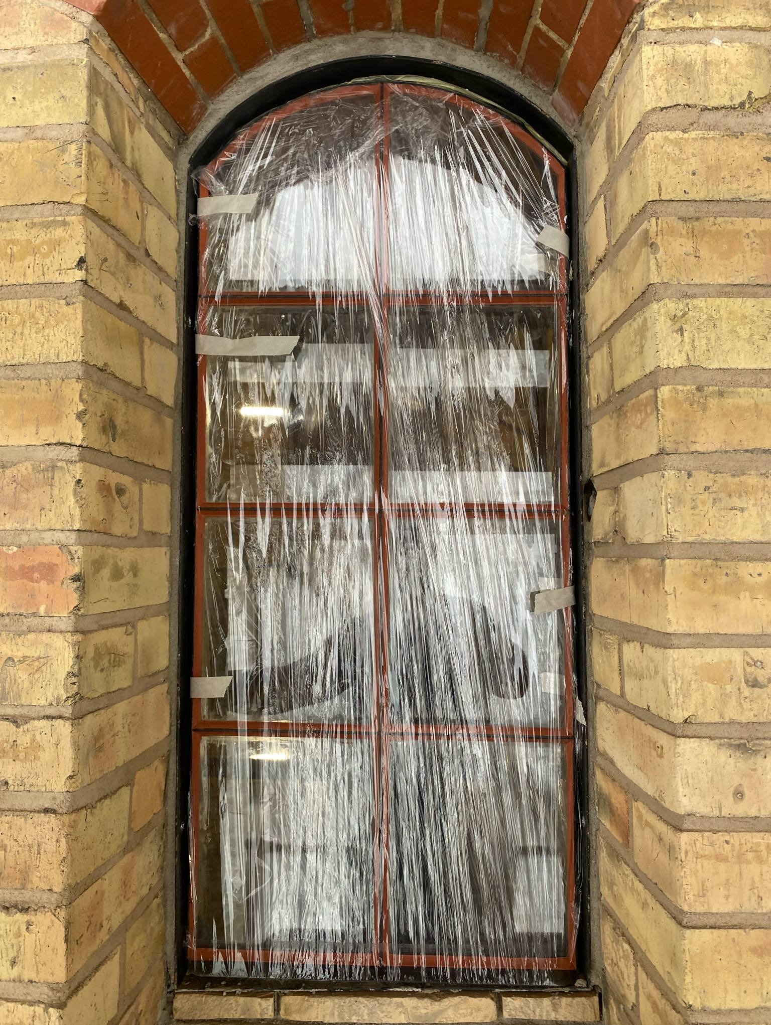 Metalowa konstrukcja okienna z łukowym nadprożem, zabezpieczona folią i taśmą, w surowym murze z cegły klinkierowej w odcieniach beżu i czerwieni.