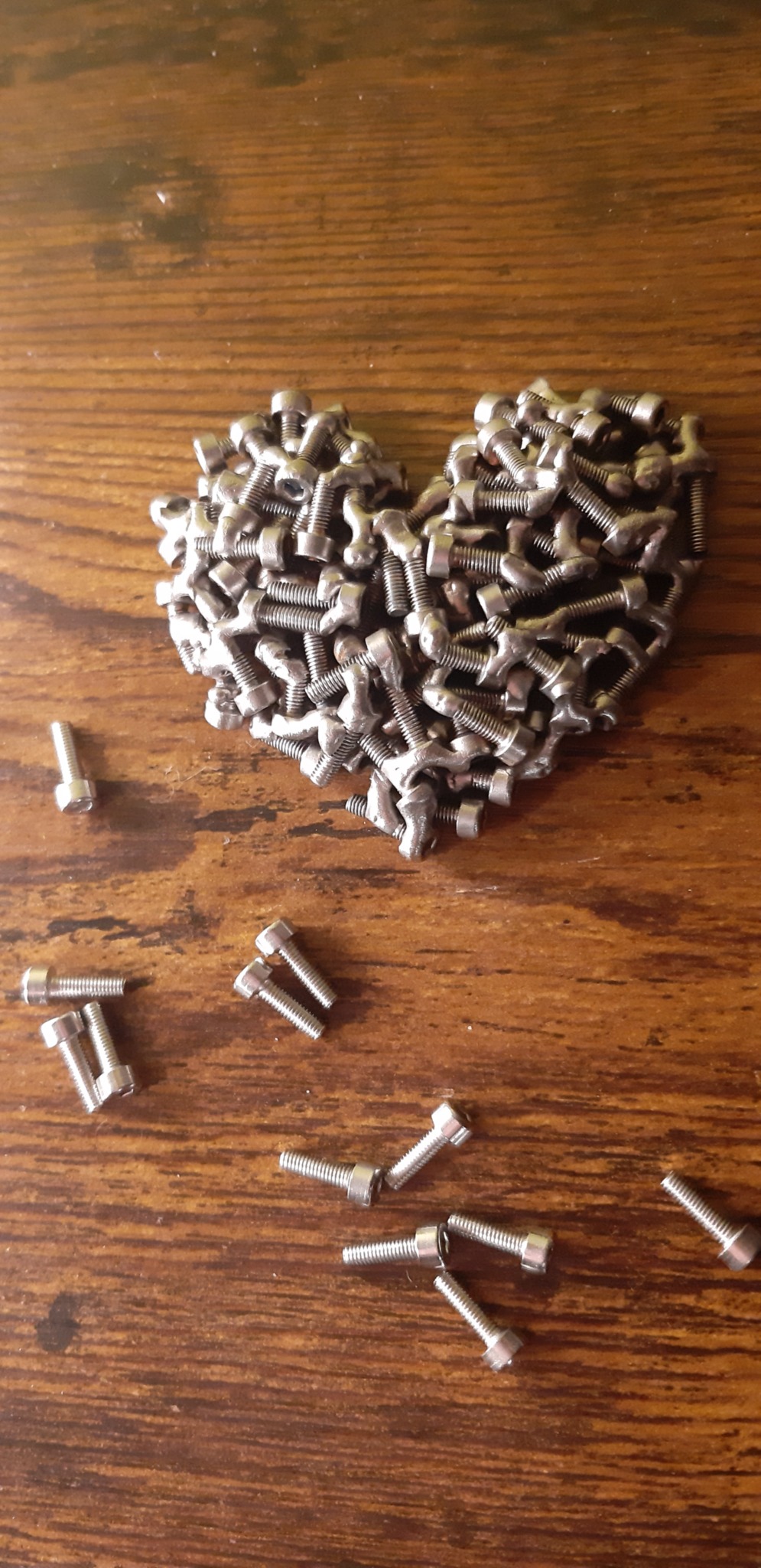 Serce ułożone ze srebrnych śrub na drewnianym tle. Kilka śrub luźno rozrzuconych wokół. Nietypowe ujęcie z góry, ukazujące metalową kompozycję.