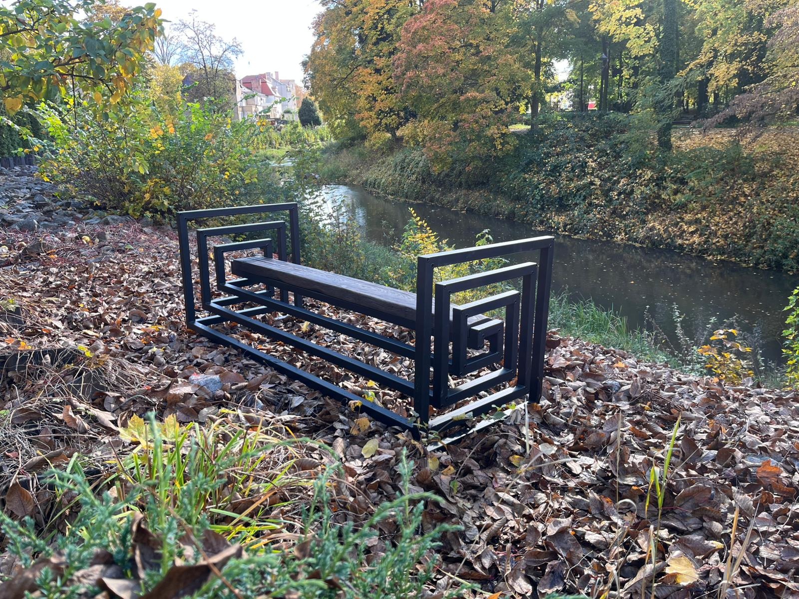 Nowoczesna, metalowa ławka parkowa z drewnianym siedziskiem, o geometrycznym designie, ustawiona na tle jesiennego krajobrazu z rzeką i drzewami.