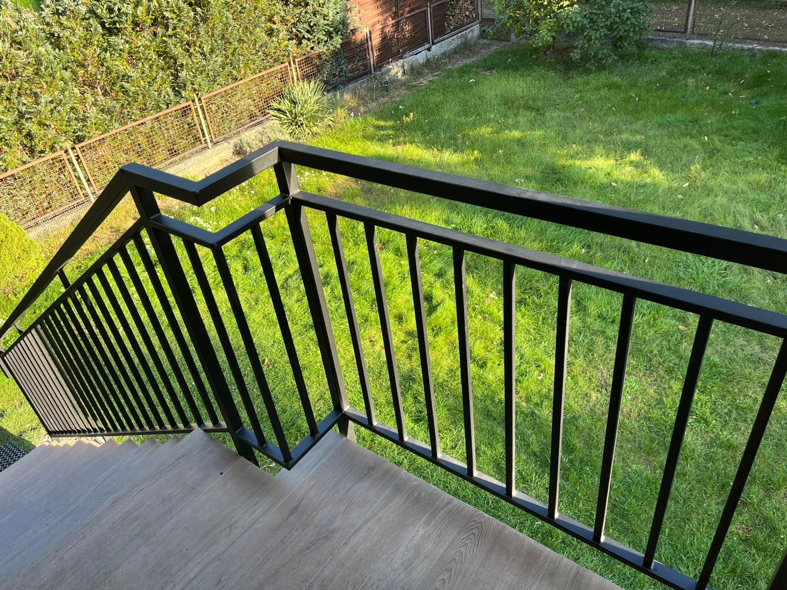 Czarna, metalowa balustrada schodowa z pionowymi szczeblami, widok z góry na schody z drewnopodobnymi panelami i zielony trawnik w tle.