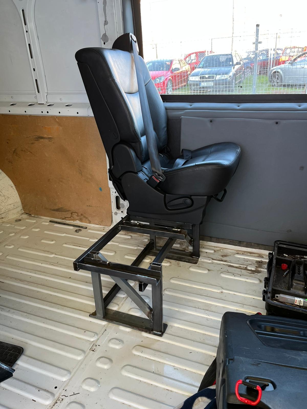 Metalowa konstrukcja pod fotel samochodowy wewnątrz pojazdu, spawana rama z profili, czarne skórzane siedzenie z pasami, narzędzia obok. Widok z boku.