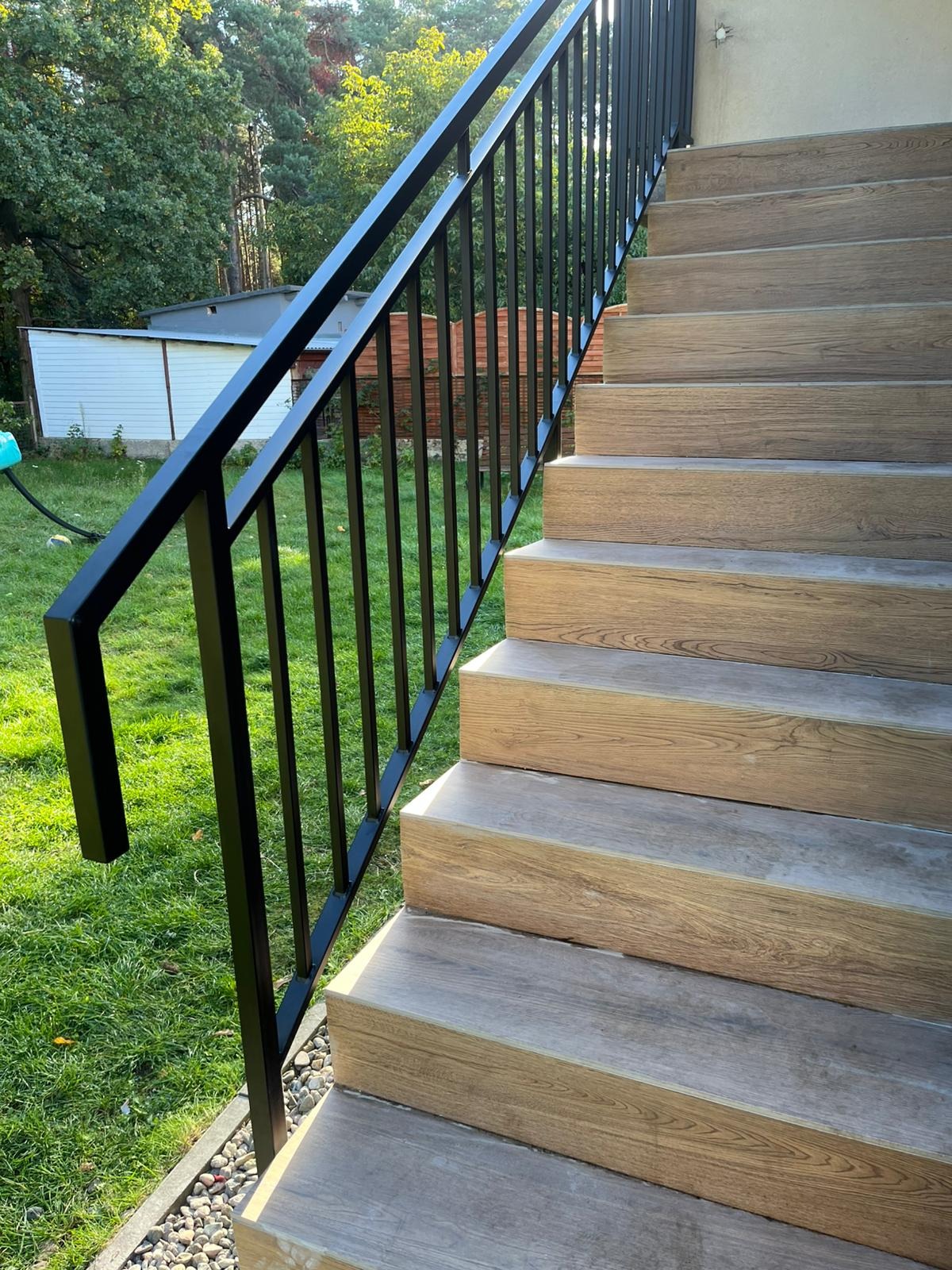 Zewnętrzne schody z drewnopodobnymi stopniami i czarną, metalową balustradą, prowadzące do budynku. Widok z dołu, zieleń w tle.