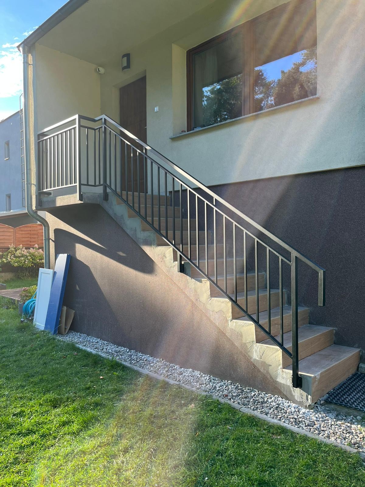 Zewnętrzne schody betonowe z czarną, metalową balustradą prowadzące do wejścia domu. Widoczne drzwi wejściowe i okno z odbiciem drzew. Na trawie oparty element w kolorze niebieskim.