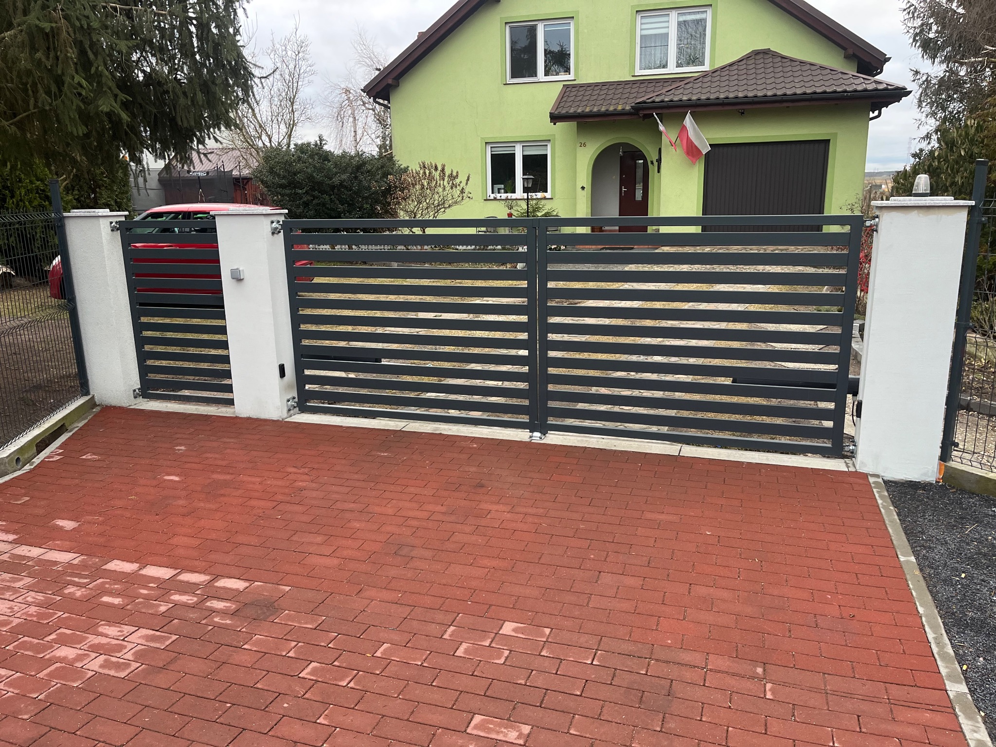 Nowoczesna, pozioma brama wjazdowa z szarego metalu, osadzona na białych słupkach. Czerwona kostka brukowa na podjeździe. Dom z zieloną elewacją w tle.