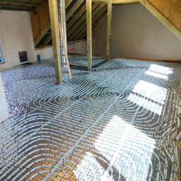 House Instal Spółka z ograniczoną odpowiedzialnością - Białe rury ogrzewania podłogowego ułożone w pętle na metalowej siatce w surowym wnętrzu poddasza, widoczne drewniane elementy konstrukcyjne dachu i ściany w trakcie wykończenia.