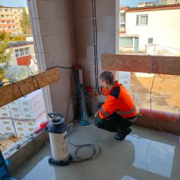 House Instal Spółka z ograniczoną odpowiedzialnością - Instalator w pomarańczowej kurtce roboczej kuca przy ścianie z pustaków, montując elementy instalacji elektrycznej w nowym budynku, z widocznym narzędziem do gięcia rur i stojącą obok pompą...