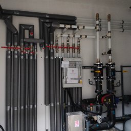 House Instal Spółka z ograniczoną odpowiedzialnością - Rozbudowana instalacja grzewcza w kotłowni: rury izolowane termicznie, zawory z czerwonymi pokrętłami, białe rury wodne, czarne pompy obiegowe Grundfos, sterownik z dokumentacją, instalacja...