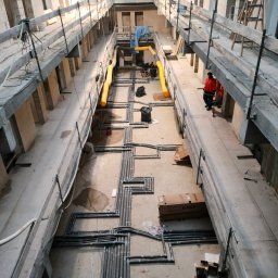 House Instal Spółka z ograniczoną odpowiedzialnością - Wysoki kąt widzenia na instalację rur w surowym wnętrzu budynku w trakcie budowy, z prowizorycznymi kładkami roboczymi i dwoma pracownikami w odblaskowych kamizelkach w oddali.