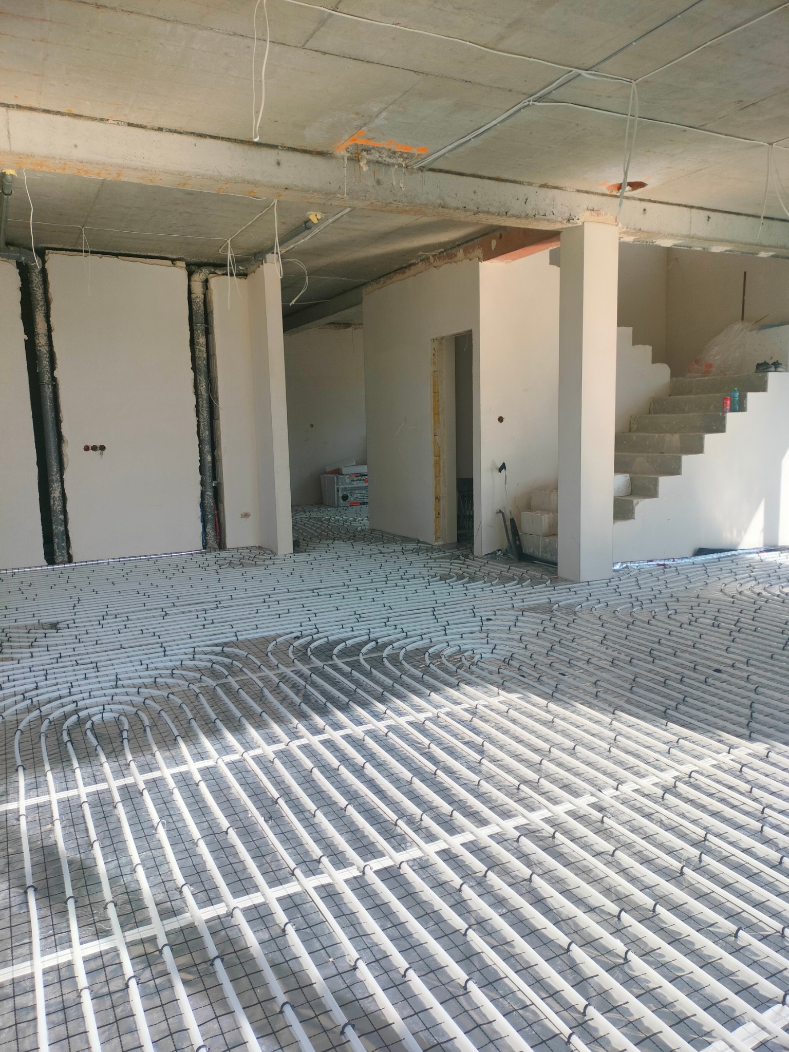 Rozłożona instalacja ogrzewania podłogowego w surowym stanie, widoczne białe rury na czarnej siatce, betonowa podłoga i ściany w trakcie budowy, schody w tle, budynek w stanie deweloperskim.