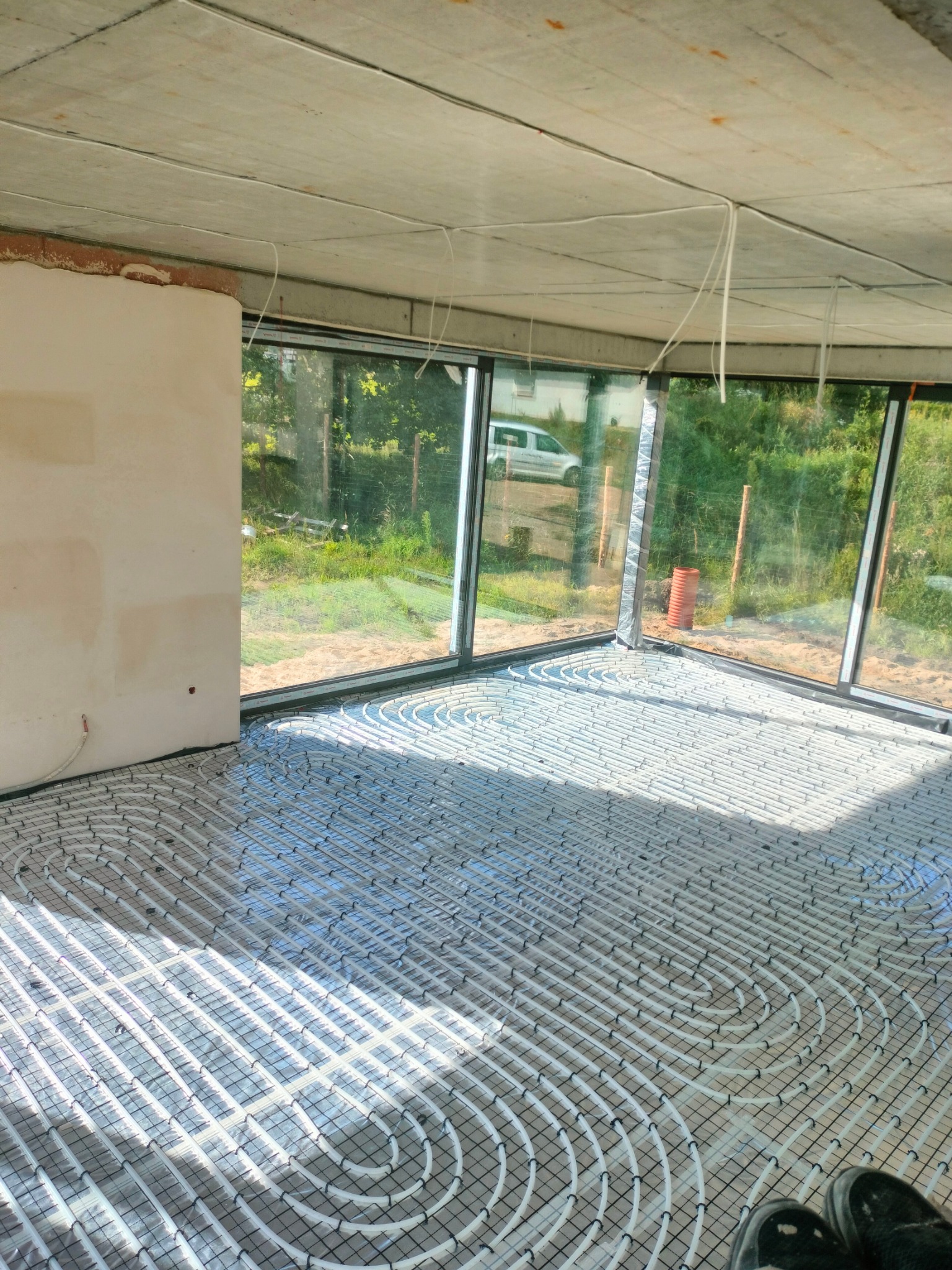 Rozłożona instalacja ogrzewania podłogowego w surowym wnętrzu z dużymi oknami, widoczne rury na czarnej siatce mocującej, betonowy sufit i fragment ściany w stanie deweloperskim.