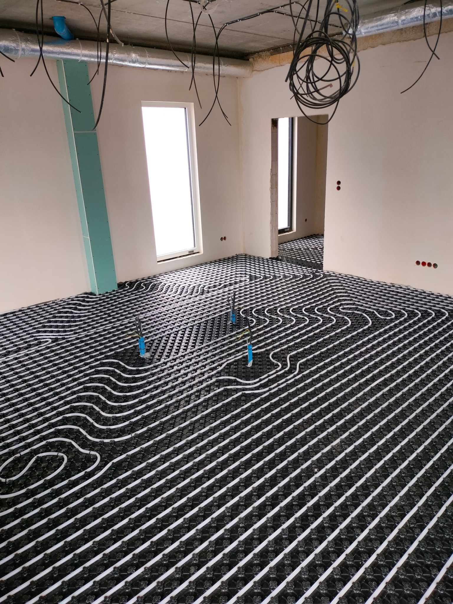 Rozległe pomieszczenie w trakcie instalacji ogrzewania podłogowego, widoczne białe rurki na czarnej macie, częściowo odsłonięte przewody elektryczne zwisają z sufitu, ściany w stanie surowym, okno...