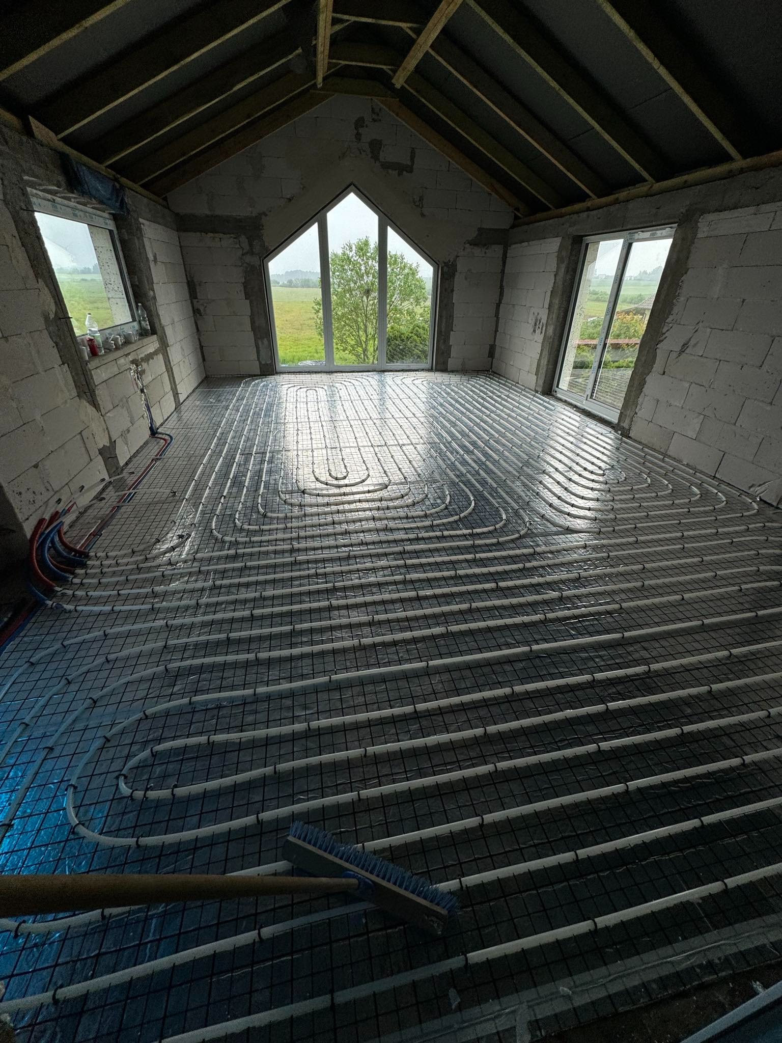 Układanie instalacji ogrzewania podłogowego w surowym wnętrzu z widokiem na krajobraz za oknem, widoczne rury grzewcze zamocowane do siatki zbrojeniowej i częściowo zalane wodą, przygotowane...