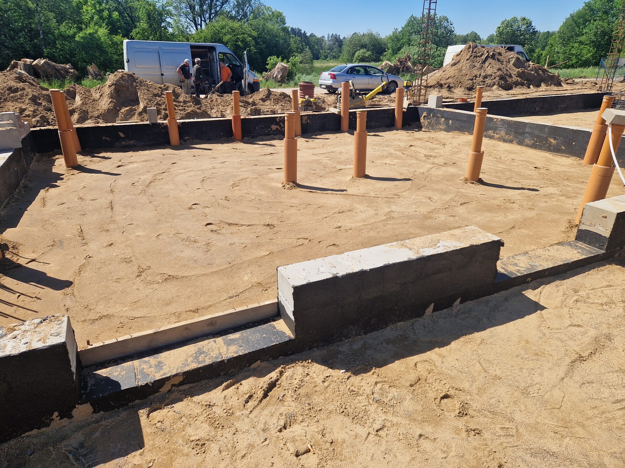 Fundament budynku w trakcie budowy z wystającymi rurami kanalizacyjnymi, widoczny samochód dostawczy i ekipa budowlana w tle.