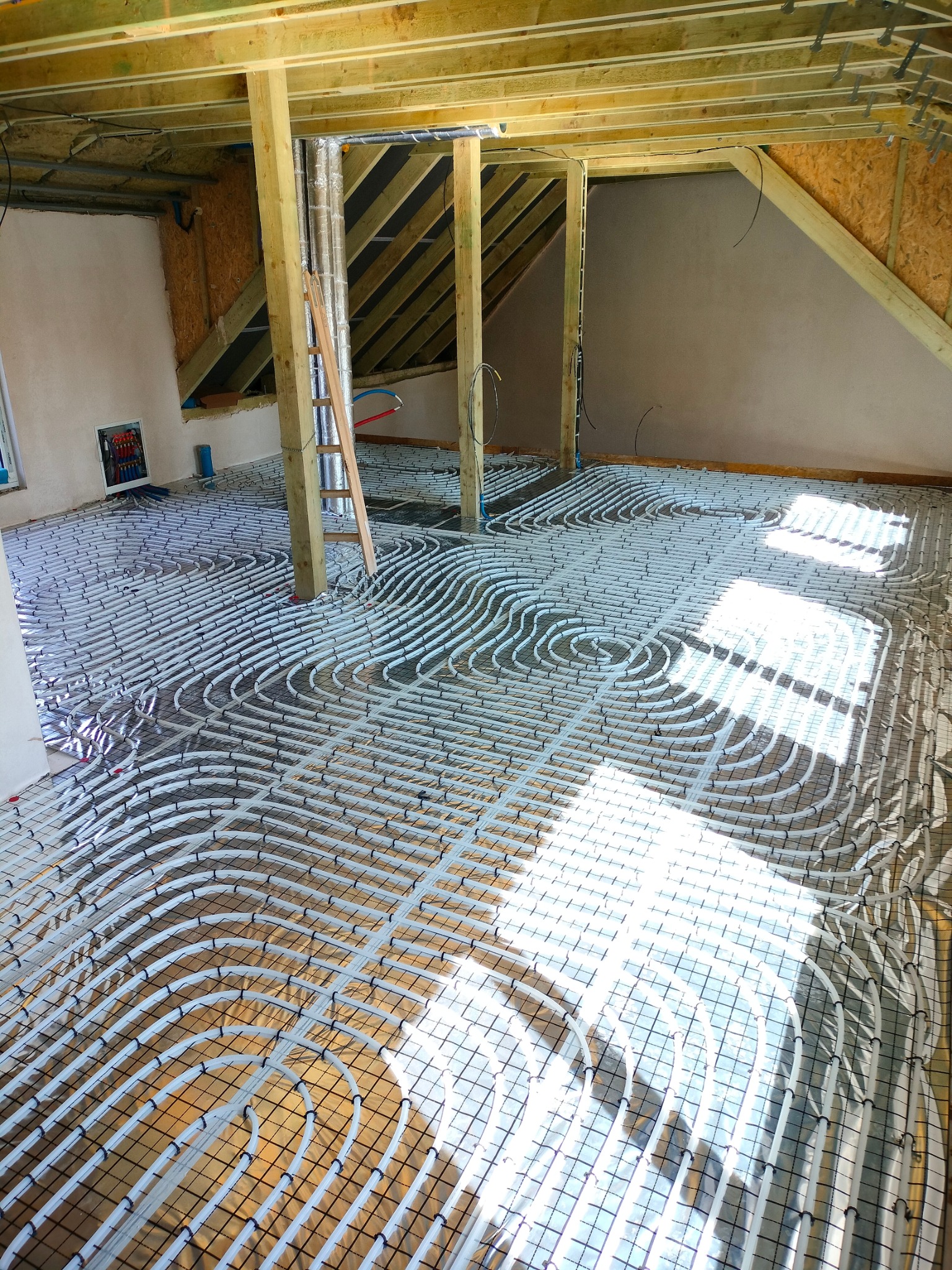 Białe rury ogrzewania podłogowego ułożone w pętle na metalowej siatce w surowym wnętrzu poddasza, widoczne drewniane elementy konstrukcyjne dachu i ściany w trakcie wykończenia.