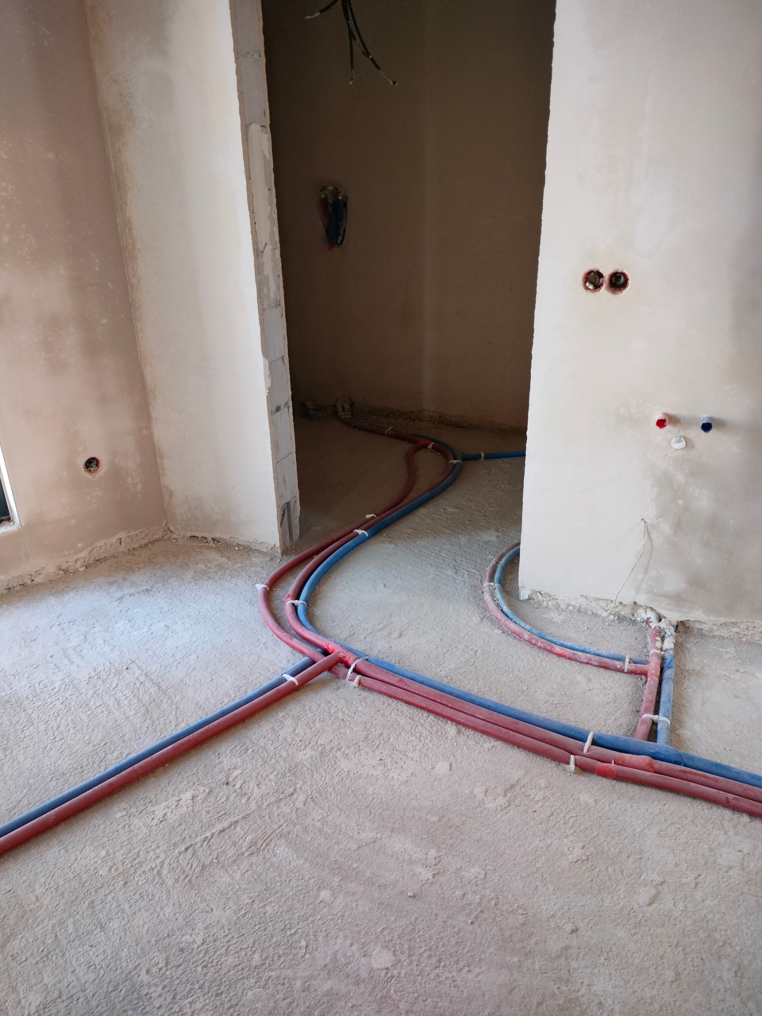 Instalacja podłogowa centralnego ogrzewania z rur PEX w kolorze czerwonym i niebieskim, zamocowanych opaskami zaciskowymi, w surowym wnętrzu podczas prac wykończeniowych.