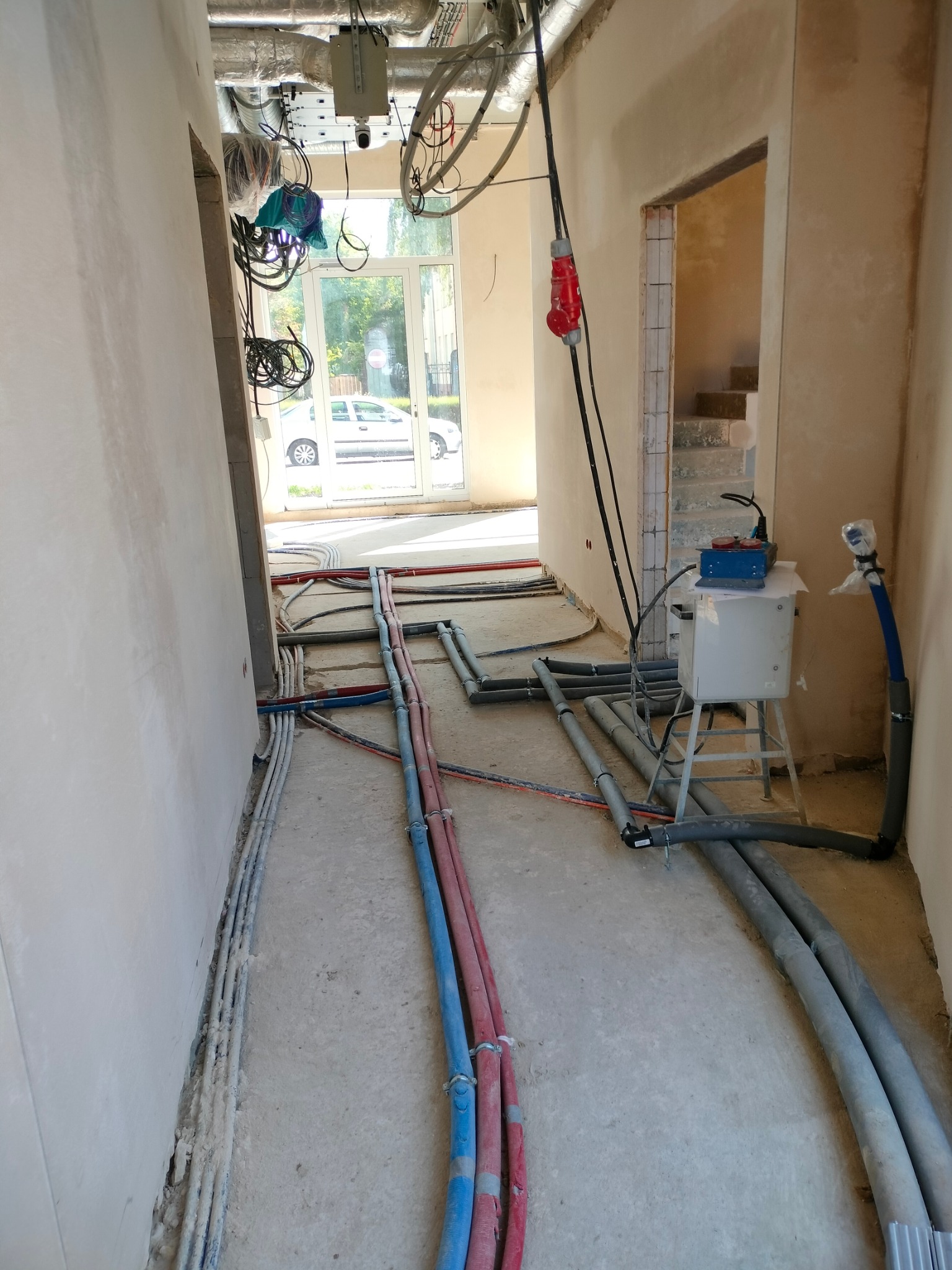 Wnętrze w trakcie prac instalacyjnych: rury grzewcze w kolorach czerwonym i niebieskim prowadzone po podłodze, szare rury kanalizacyjne, przewody elektryczne przy ścianie, oraz urządzenie pomiarowe...