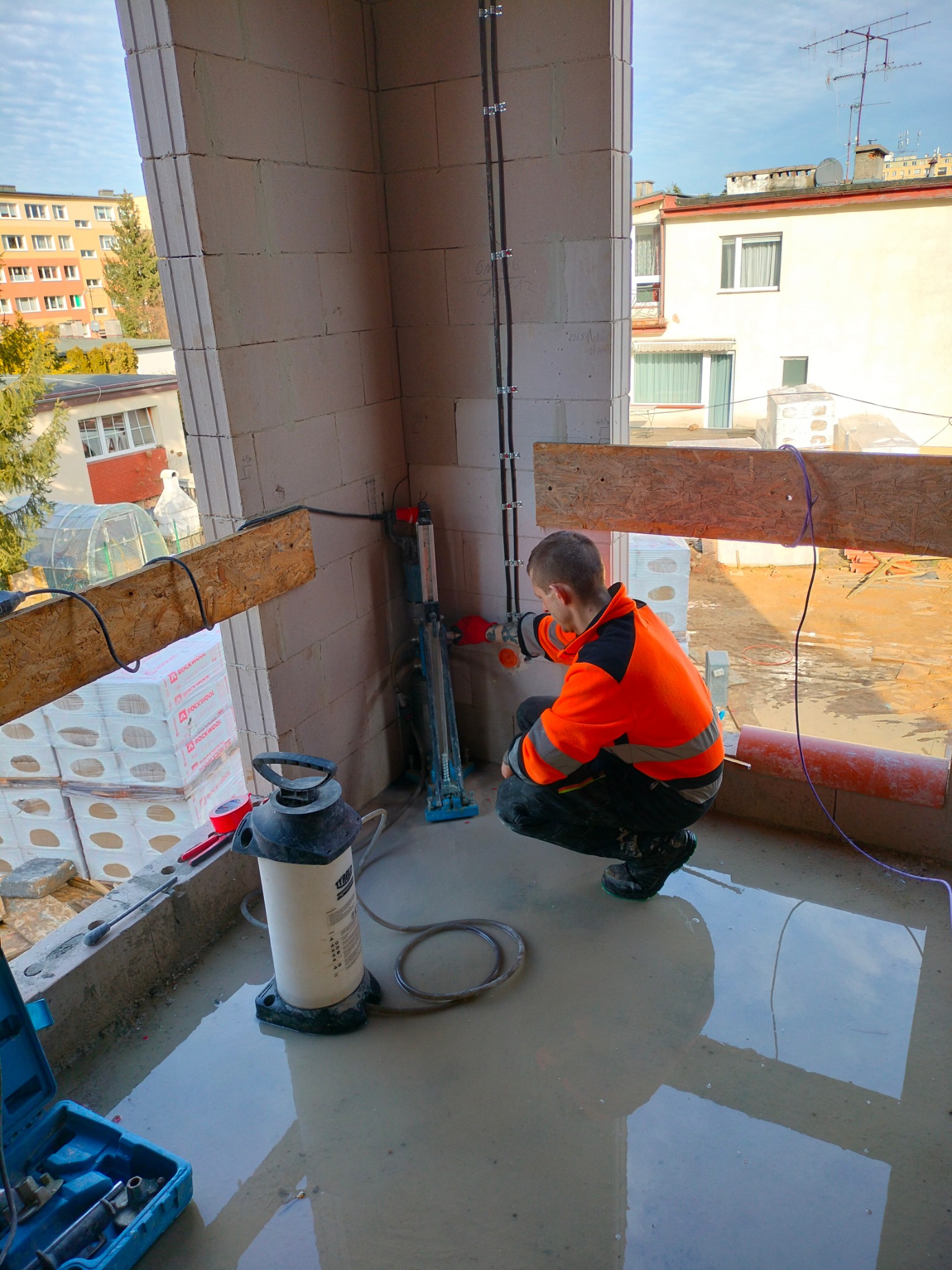 Instalator w pomarańczowej kurtce roboczej kuca przy ścianie z pustaków, montując elementy instalacji elektrycznej w nowym budynku, z widocznym narzędziem do gięcia rur i stojącą obok pompą...