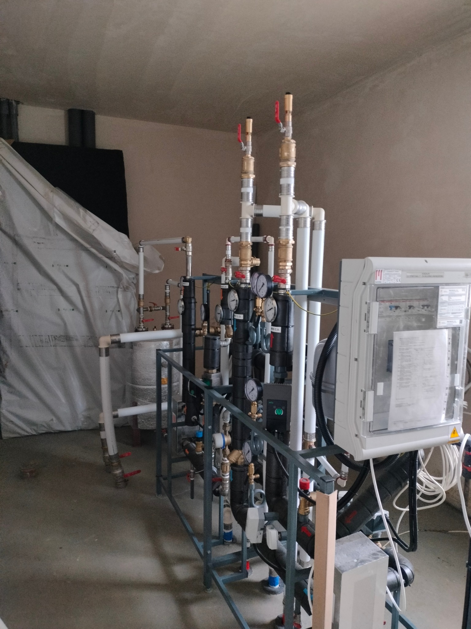 Rozbudowana instalacja grzewcza z licznymi rurami, zaworami i manometrami, osadzona na metalowej konstrukcji. Na pierwszym planie biała skrzynka sterownicza z dokumentacją. W tle zbiornik owinięty...