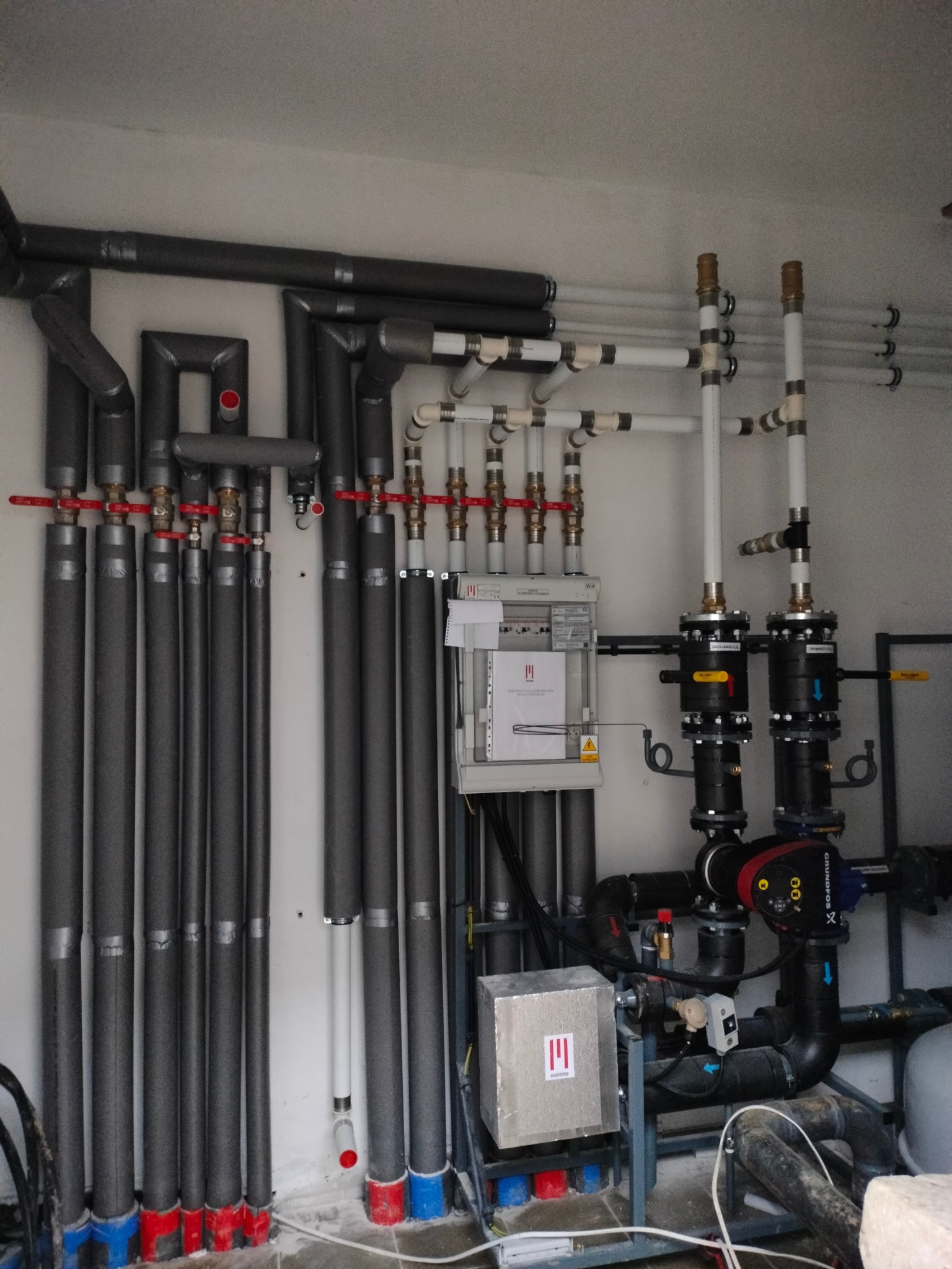 Rozbudowana instalacja grzewcza w kotłowni: rury izolowane termicznie, zawory z czerwonymi pokrętłami, białe rury wodne, czarne pompy obiegowe Grundfos, sterownik z dokumentacją, instalacja...