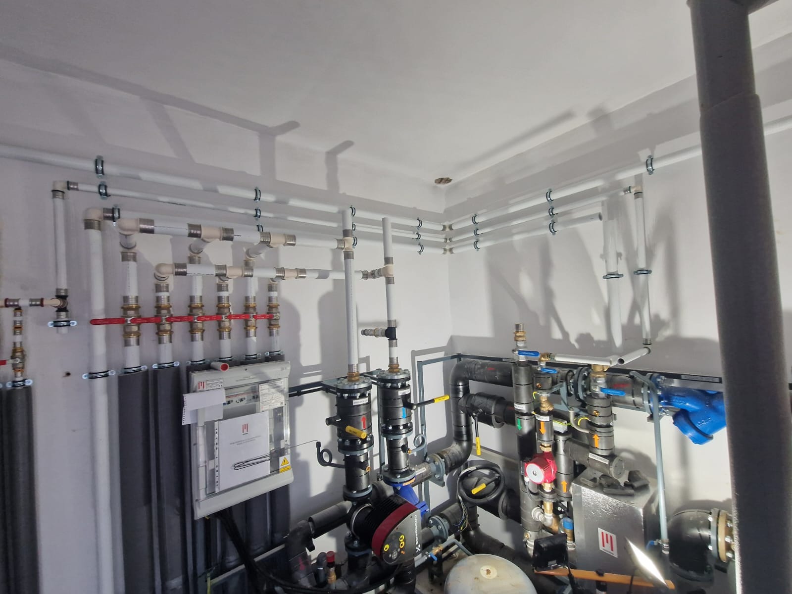 Rozbudowana instalacja grzewcza z rurami PCV i metalowymi elementami, widoczne zawory, pompy obiegowe i tablica sterownicza na ścianie, całość umieszczona w pomieszczeniu technicznym.