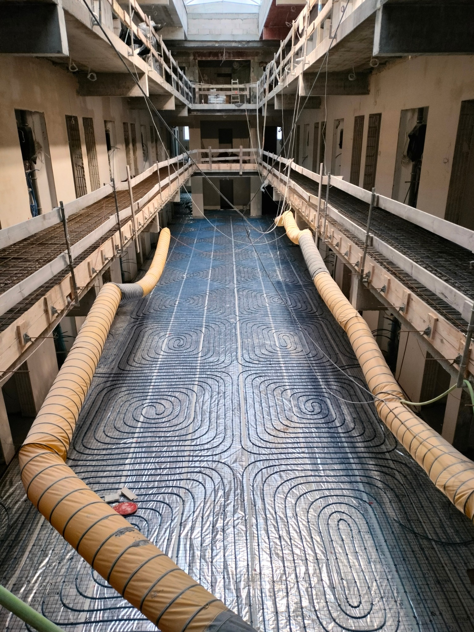 Widok z góry na rozłożoną instalację ogrzewania podłogowego w surowym stanie, z żółtymi rurami wentylacyjnymi po bokach, wewnątrz budynku w trakcie budowy.