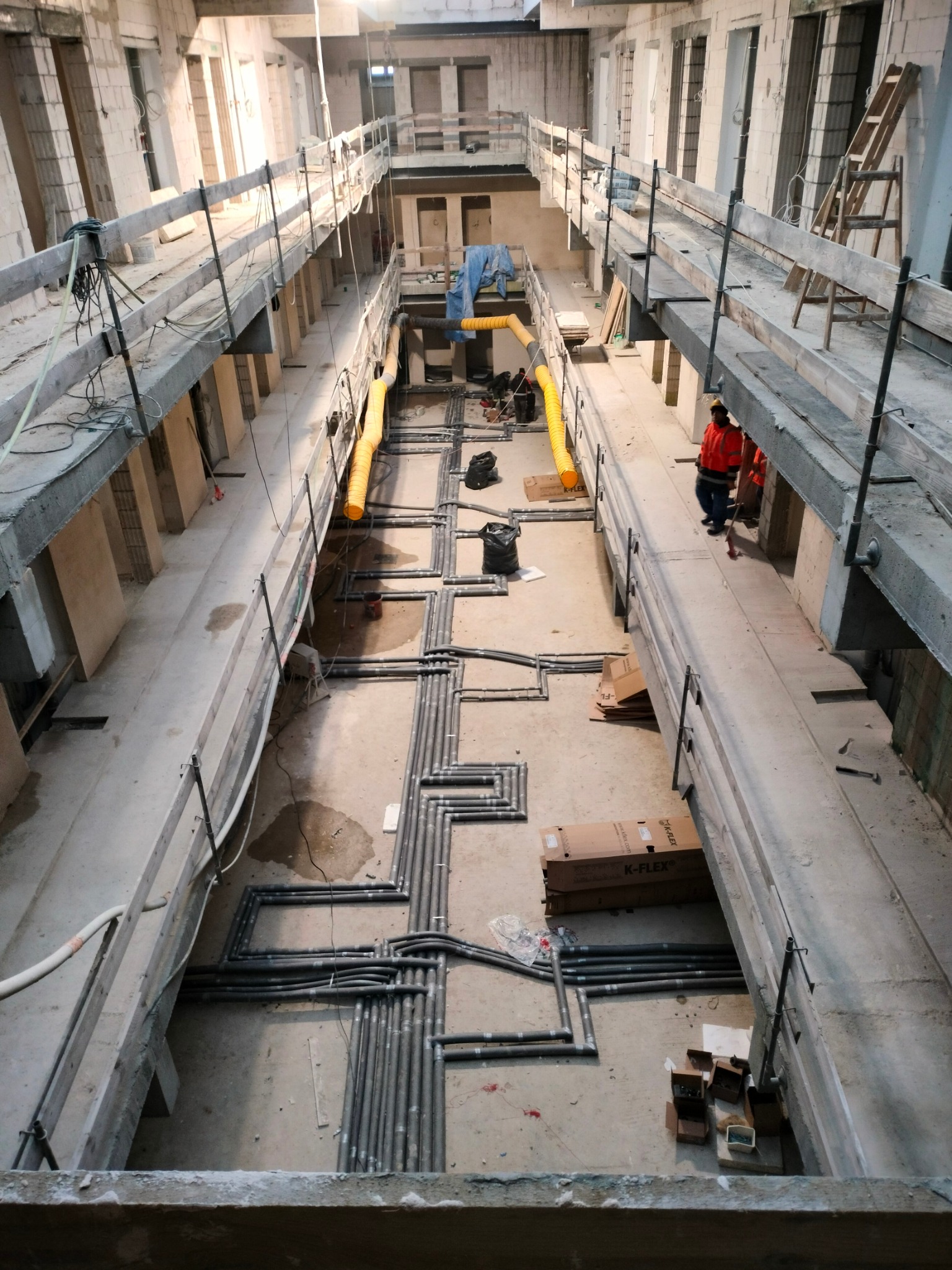 Wysoki kąt widzenia na instalację rur w surowym wnętrzu budynku w trakcie budowy, z prowizorycznymi kładkami roboczymi i dwoma pracownikami w odblaskowych kamizelkach w oddali.