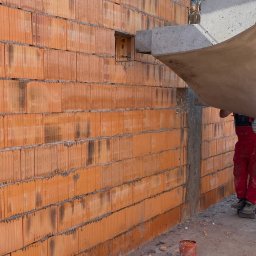 HRO SPÓŁKA Z OGRANICZONĄ ODPOWIEDZIALNOŚCIĄ - Wznoszenie ściany z pustaków ceramicznych; widoczny otwór okienny, betonowy nadproże podnoszone dźwigiem oraz pracownik w czerwonym kombinezonie.