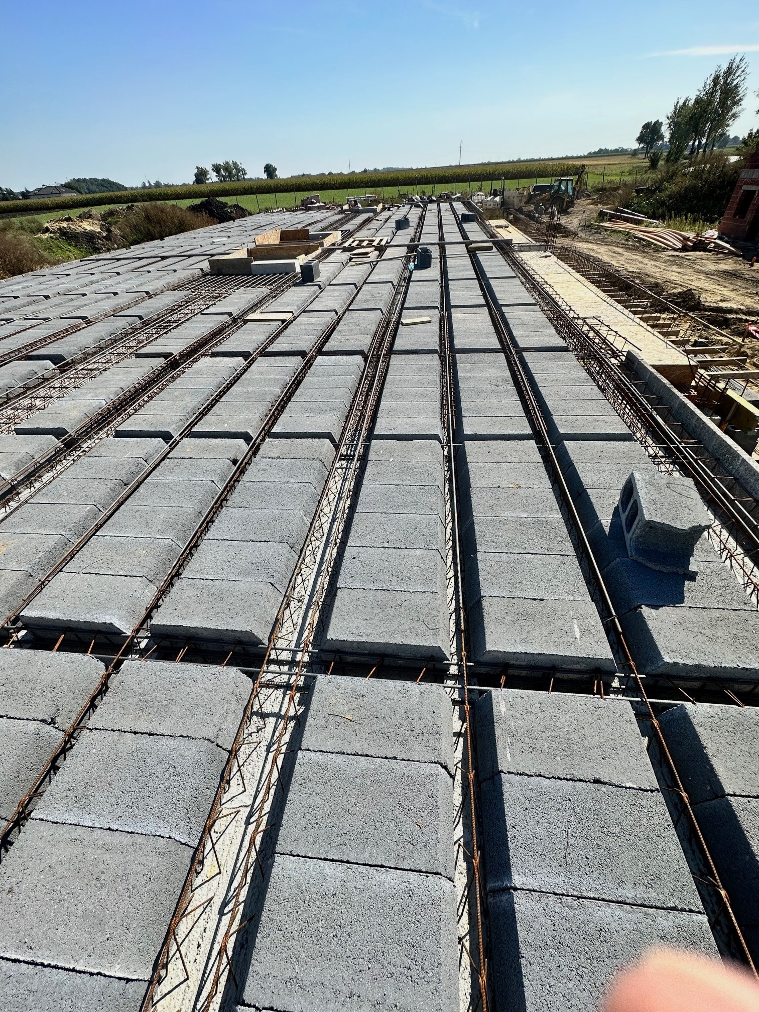 Wiązanie zbrojenia na stropie Teriva z szarych betonowych pustaków, widok z góry, w tle krajobraz polnych upraw.