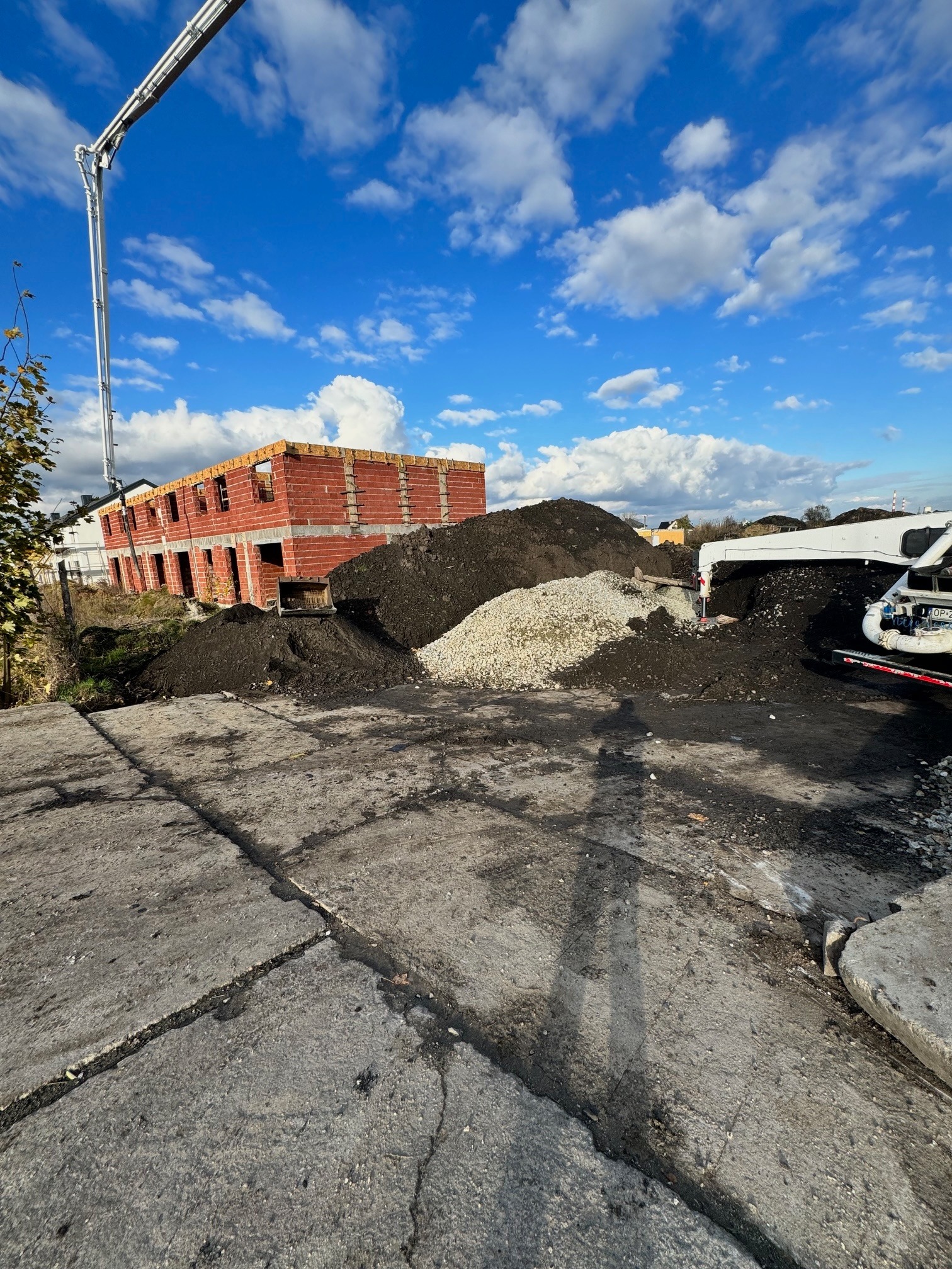 Dwukondygnacyjny dom murowany w trakcie budowy, widoczny czerwony mur z pustaków, obok hałda ziemi i żwiru, w tle błękitne niebo z chmurami i ramię pompy do betonu.