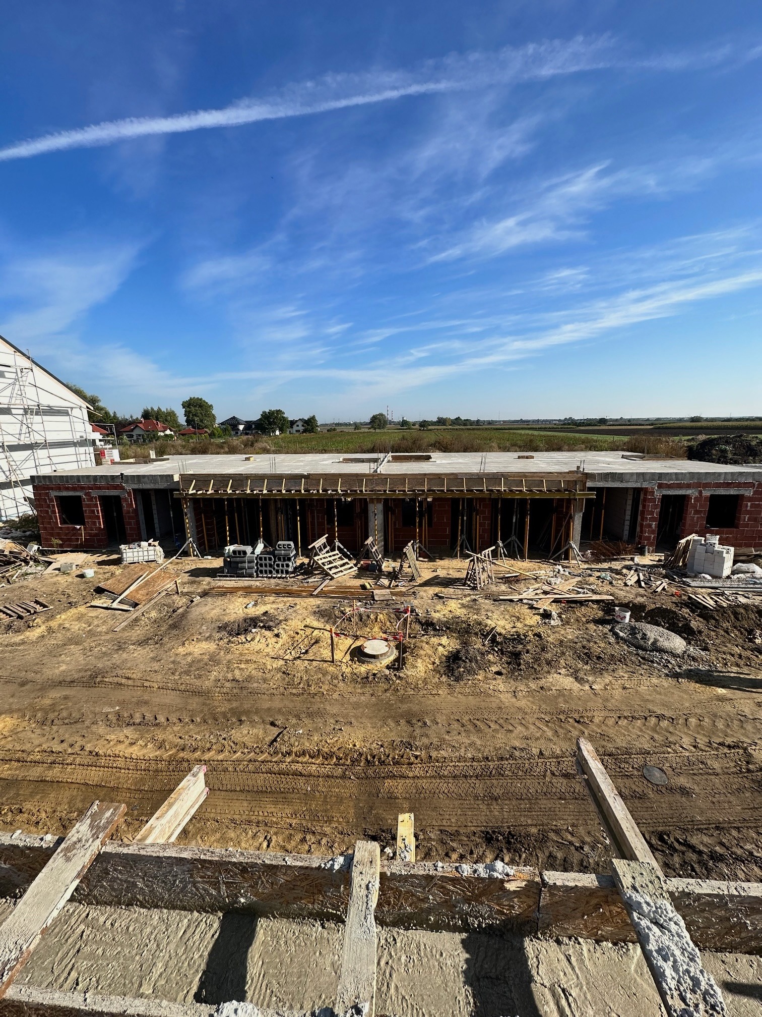 Budowa parterowego domu w stanie surowym otwartym z czerwonej cegły, widoczne szalunki stropu, na zewnątrz plac budowy z widocznymi śladami opon, w tle zabudowa jednorodzinna i błękitne niebo...