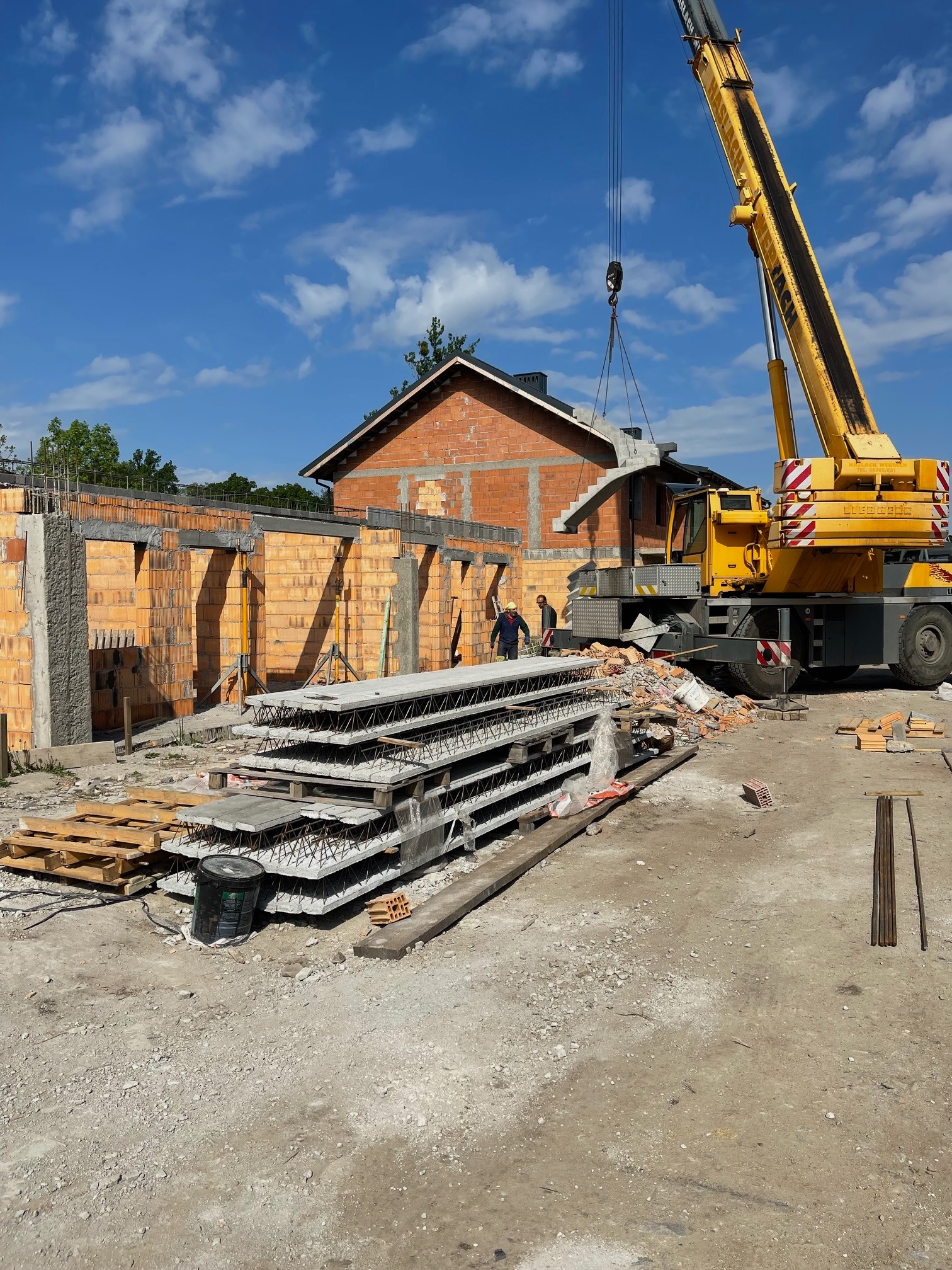Żuraw budowlany podnosi prefabrykowane betonowe schody na budowę domu z czerwonej cegły, widoczni dwaj pracownicy w oddali, na pierwszym planie stos płyt betonowych i palety.
