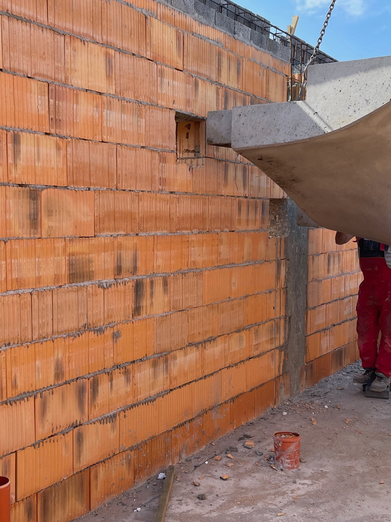Wznoszenie ściany z pustaków ceramicznych; widoczny otwór okienny, betonowy nadproże podnoszone dźwigiem oraz pracownik w czerwonym kombinezonie.
