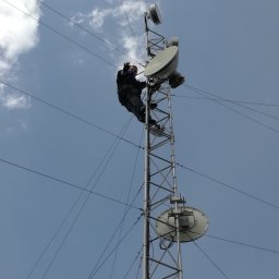 LanSystem Paweł Sutyła - Pracownik na szczycie wieży telekomunikacyjnej, instaluje antenę satelitarną na tle błękitnego nieba z chmurami. Ujęcie z dołu podkreśla wysokość konstrukcji.