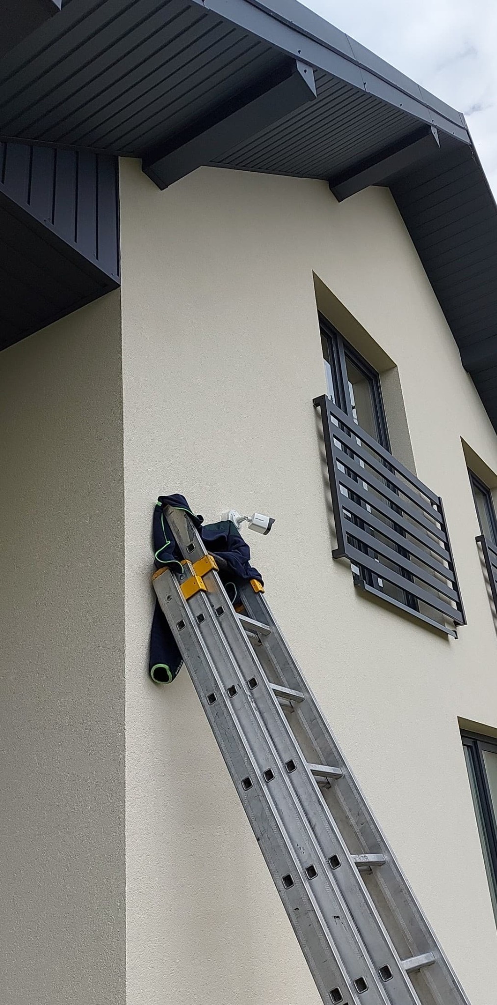 Montaż kamery na elewacji domu z drabiny. Widoczna kamera, drabina oparta o ścianę, fragment budynku z oknami i szarą elewacją. Ujęcie z dołu.